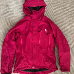 Rosa vindjacka från Haglöfs S - Snygg rosa vindjacka från Haglöfs med huva, flera dragkedjeförsedda fickor och justerbara detaljer. Jackan är i funktionsmaterial och har en sportig look, perfekt för dig som gillar att vara ute oavsett väder. Märke synligt på bröstet.