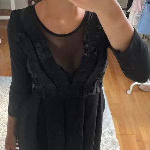 Svart klänning med mesh - Svart kortklänning från H&M med trekvartsärmar och snygga plisserade detaljer framtill. Djup v-ringning med transparent meshpanel ger en cool look. Kjolen är lätt utställd och materialet känns mjukt och följsamt.
