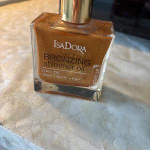 Isadora Bronzing Shimmer Oil Gold - Isadora Bronzing Shimmer Oil i nyansen 61 Gold. En torr olja med intensivt guldig lyster som ger huden och håret en snygg glow. Kommer i en fyrkantig glasflaska med guldfärgad kork. Passar både ansikte, kropp och hår för en solkysst look.
