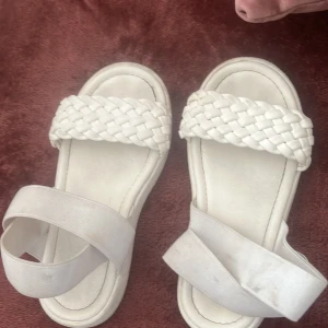 Vita flätade sandaler med resårband - Snygga vita sandaler med bred flätad rem över tårna och elastiskt band runt hälen för extra komfort. Platt sula och stilren design som passar perfekt till sommarens outfits. Gjorda i syntetmaterial med en modern look.