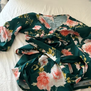 Blommig grön bluese från Nelly - Supersöt grön bluese från Nelly med stora rosa och vita blommor. Omlottmodell med v-ringning och långa ärmar. Perfekt för sommarens alla tillfällen och ger en romantisk vibe. Sval och lätt i materialet.