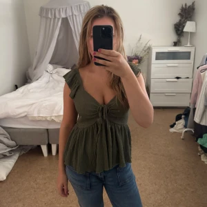 Olivgrön blus med volangärm och knyt - Supersöt olivgrön blus med volangärmar och knyt framtill. Toppen har en lätt rynkad struktur och v-ringning, samt en flowy passform nedtill. Perfekt till jeans eller kjol, använt en gång