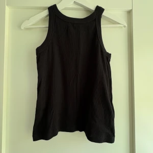 Svart ribbad linnetopp - En svart ribbad linnetopp med klassisk rund halsringning och breda axelband. Toppen är ärmlös och har en enkel, stilren design som passar perfekt till jeans eller kjol. Materialet är mjukt och stretchigt för en bekväm passform.