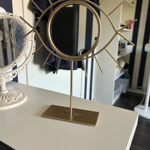 Guldigt öga-formad smyckesställ - Snyggt smyckesställ i form av ett öga i guldig metall. Perfekt för att hänga ringar, armband eller halsband. Stället har en rund spegel i mitten och stilrena linjer som ger en modern vibe till rummet. Köpt på Jysk 