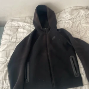 Svart Nike hoodiejacka XL - Svart hoodiejacka från Nike i storlek XL. Jackan har hel dragkedja framtill, två fickor med dragkedja och en stilren Nike-logga på bröstet. Mjuk och skön, perfekt för chill eller träning. Klassisk passform med huva och långa ärmar.