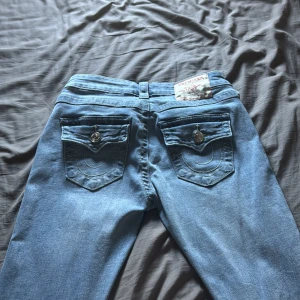 True Religion bootcut jeans ljusblå - Säljer ett par ikoniska ljusblå jeans från True Religion med snygga detaljerade bakfickor och silverfärgade knappar. Jeansen har bootcut-modell och klassisk femficksdesign. Perfekta för dig som vill ha en cool och tidlös denimlook.