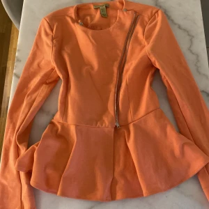 Persikofärgad jacka från Only Trend - Snygg persikofärgad jacka från Only Trend med lång ärm och cool sned dragkedja framtill. Jackan har en peplum-liknande nederdel som ger en feminin siluett. Perfekt för dig som vill sticka ut med färg och form! Finns två små fläckar 