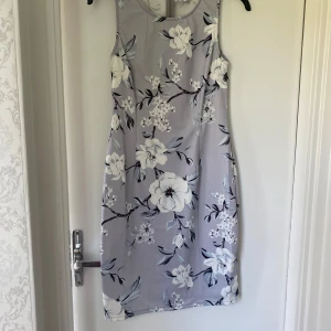 Grå  blommig ärmlös klänning - Superfin grå kortklänning med stora vita och grå blommor. Ärmlös modell med figurnära passform och dragkedja i ryggen. Perfekt för dig som gillar romantiska och stilrena vibes. Materialet känns mjukt och följsamt.