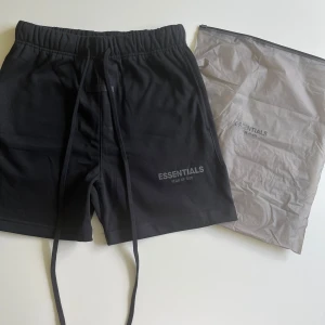 Svarta Essentials shorts med snörning - Svarta shorts från Essentials med vit logga på vänster ben. De har elastisk midja med snörning och en bakficka. Perfekta för chill dagar eller träning, med en loose fit och mjukt material som känns skönt mot huden.