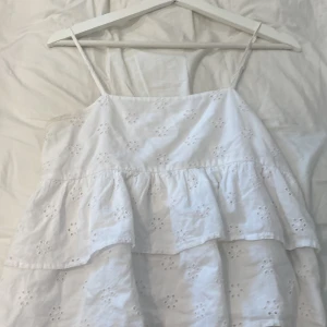 Vit volangtop från Gina Tricot - Superfin vit linnetopp från Gina Tricot med tunna axelband och broderade blommor. Toppen har dubbla volanger och är luftig, perfekt för varma dagar. Söt och enkel att matcha med jeans eller kjol.