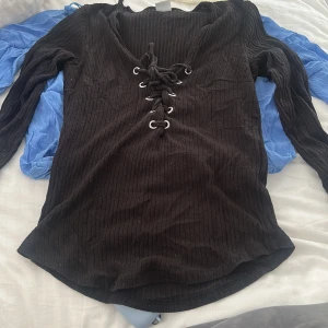 Svart ribbad tröja med snörning Gina Tricot - Svart långärmad tröja från Gina Tricot med ribbad struktur och snygg snörning framtill. Tröjan har v-ringning och är figurnära i modellen. Den är lite sönder i nacken som man ser på den andra bilden