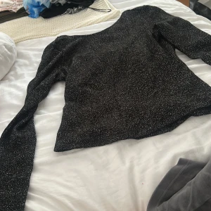 Glittrig svart långärmad topp - Säljer en svart långärmad topp med glittriga inslag. Toppen har en rund halsringning och är figurnära i modellen. 