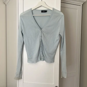 Ljusblå kofta från Mexx - Supersöt ljusblå kofta från Mexx med v-ringning och knappar framtill. Den är ribbad vid ärmslut och nederkant, har lång ärm och är i ett tunt, mjukt material som passar perfekt till lager-på-lager. Enkel och stilren design som funkar till det mesta.