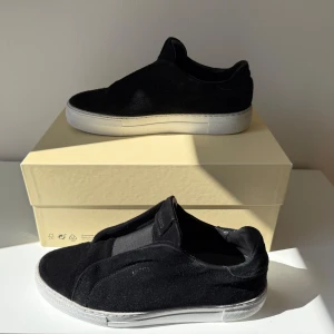 Axel Arigato Clean 360 - Stilrena svarta sneakers från Axel Arigato i mocka med vit platt sula. Skorna har minimalistisk design utan snörning och diskret logga på sidan. Perfekta för dig som gillar en clean och modern look. (Allt original medföljer)