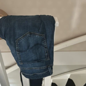Mörkblå jeans från Jack and Jones  - Säljer ett par mörkblå jeans från Jack and Jones  med klassisk femficksdesign och snygga slitningar. Jeansen har raka ben och normal passform, tillverkade i mjukt bomullsjeansmaterial. Perfekta för en avslappnad och stilren look. Använd 1 gång 