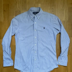 Ralph lauren skjorta - Säljer denna riktigt snygga randiga Ralph lauren skjorta i vitt och blått, den är i mycket bra skick, storlek S modell custom fit den är 73 cm lång och 48 cm bred under armhålorna, hör gärna av dig vid frågor!