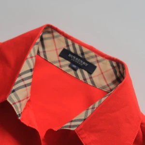 Röd skjorta från Burberry med rutigt foder - Snygg röd skjorta från Burberry med klassiskt rutigt foder i kragen. Skjortan har lång ärm, knappar framtill och en figurnära passform. Perfekt för dig som vill sticka ut med en ikonisk detalj från ett lyxigt märke.