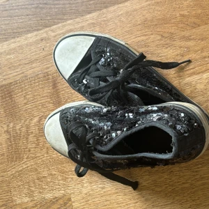 Svarta sneakers med paljetter - Coola svarta sneakers med glittriga paljetter över hela skon och vita tåpartier. Skorna har svarta snören och en klassisk platt sula. Perfekta för dig som vill sticka ut med lite extra bling i vardagen.