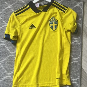 Svenska landslagströja Adidas gul - Gul fotbollströja från Adidas med Sveriges landslagsemblem på bröstet och svarta detaljer. Tröjan har korta ärmar, svart krage och klassiska Adidas-ränder på axlarna. Tillverkad i lätt och ventilerande AEROREADY-material, perfekt för match eller träning. Ändå är att det står Albin i den, men det syns inte när man har på den