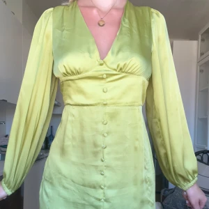 Grön kort klänning med knappar - Superfin grön kort klänning i glansigt material med v-ringning och långa ärmar. Klänningen har markerad midja och dekorativa knappar framtill hela vägen ner. Perfekt för dig som vill sticka ut med en färgstark look. Storlek 40 men känns som en M