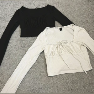 Svart och vit croppad långärmad topp - Två croppade långärmade toppar i svart och vitt. Båda har fyrkantig urringning och rynkade detaljer framtill, den vita har även en söt knytning vid bysten. Skönt och mjukt material- inte jätte tjockt. Lite för korta för mig men perfekt för dig som köper!