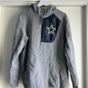 Grå Dallas Cowboys hoodie från Nike - Snygg grå hoodie från Nike med Dallas Cowboys-stjärnan på bröstet. Tröjan har huva med dragsko, lång ärm och en marinblå panel med dragkedja framtill. Tillverkad i mjuk bomullsblandning, perfekt för NFL-fans som vill sticka ut. Importerat från USA. 