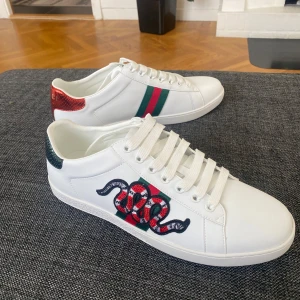Gucci Ace skor - Snygga vita Gucci Ace sneakers i skinn med klassiska gröna och röda ränder på sidan samt broderad orm. Baksidan har metallicröd och metallicgrön detalj med ormskinnsmönster och Gucci-logga. Perfekta för dig som vill sticka ut med lyxig streetstyle.