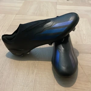Adidas X Crazyfast svarta/blå fotbollsskor - Snygga Adidas X Crazyfast fotbollsskor i svart med blå metallic-detaljer och klassiska tre ränder på sidan. Skorna har en strömlinjeformad design utan snörning och är tillverkade i syntetmaterial för lätt känsla och maxad fart på planen.