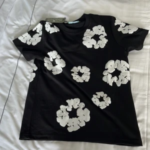 Svart blommig t-shirt från Denim Tears - Svart t-shirt från Denim Tears med stora vita blommiga tryck över hela plagget. Klassisk passform med rund hals och korta ärmar. Tillverkad i mjuk bomull, perfekt för en cool och avslappnad stil. (Köptes idag men den var för stor för mig)