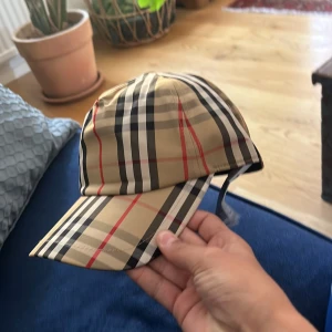 Burberry rutig keps beige M - Snygg keps från Burberry i klassiskt beige rutigt mönster med svarta, vita och röda linjer. Justerbar baktill med svart spänne och tydlig Burberry London England-logga. Tillverkad i polyester, perfekt för att lyfta din outfit.