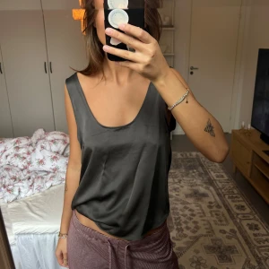 Mörkgrå siden-linnetopp - Snygg mörkgrå linnetopp i glansigt sidenmaterial med djup rundad urringning. Toppen är ärmlös och har en avslappnad passform som faller snyggt mot kroppen. Perfekt att styla med jeans eller kjol för en trendig look.