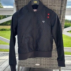 Svart bomberjacka Fred Perry x Amy Winehouse - Svart sommarjacka från Fred Perry x Amy Winehouse Foundation med diskret rutigt mönster, röda hjärtan på bröstet och klassisk Fred Perry-logga. Jackan har dragkedja, ribbade muddar och två snedställda fickor. Perfekt för dig som vill ha en unik och stilren look.