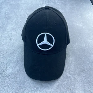 Svart Mercedes keps med broderad logga - Svart keps från Mercedes-Benz med den klassiska silverbroderade loggan framtill. Justerbar rem baktill med metallspänne som också har Mercedes-emblemet. Kepsen är i bomull och har en klassisk böjd skärm. Perfekt accessoar för bilentusiasten.