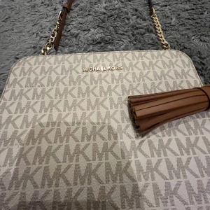 Beige axelväska Michael Kors med tofs - Snygg beige axelväska från Michael Kors med MK-logga över hela väskan. Väskan har guldfärgade kedjedetaljer, justerbar axelrem i brunt och en stor brun tofs som dekoration. Perfekt för dig som gillar stilrena och trendiga accessoarer. Pris kan diskuteras.
