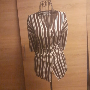 Randig blus från Lindex - Snygg blus från Lindex med bruna och vita vertikala ränder. Klänningen har v-ringning, knappar framtill och trekvartsärm. Perfekt för dig som gillar en stilren och trendig look. Materialet känns lätt och skönt mot huden.