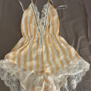 Randig peignoir med spets - Supersöt peignoir och vit randig pejgnoir i satin med djup v-ringning och tunna axelband. Toppen har breda spetsdetaljer längs urringningen och nederkanten, vilket ger en romantisk vibe. Perfekt för dig som gillar mjuka material och snygga detaljer.