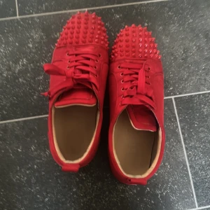 Röda Christian Louboutin sneakers med nitar - Unika röda sneakers från Christian Louboutin med coola nitar över tån och matchande röd sula med logga. Skorna har snörning och är tillverkade i skinn och mocka för en lyxig känsla. Perfekta för dig som vill sticka ut med en edgy vibe.