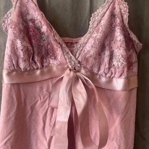 Rosa spetsig linnetopp med rosett - Supersöt ljusrosa linnetopp med spetsdetaljer och blommigt mönster upptill.🩷🩷 Satinband under bysten som knyts till en rosett framtill. Axelbanden är också i spets och toppen är ärmlös. Perfekt för dig som gillar romantisk och feminin stil.💖💖💖😜😜