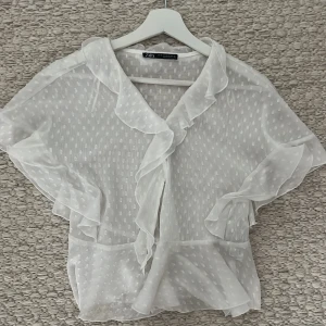 Vit volangblus från Zara XS - Superfin vit blus från Zara i transparent, lätt material med diskret prickigt mönster. Blusen har volangdetaljer längs krage och ärmslut samt korta ärmar. Helt nyskick!