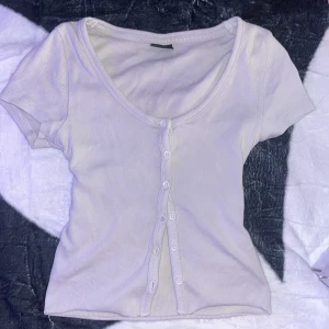 Vit ribbad topp med knappar - Superfin vit ribbad topp med korta ärmar och knäppning hela vägen framtill. Djup rundad urringning och figurnära passform gör den perfekt till jeans eller kjol. Enkel och stilren design som passar till många olika looks.