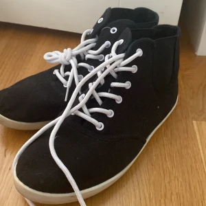 Svarta höga sneakers med vita snören - Svarta sneakers i canvas med högt skaft och vita snören. Skorna har vit sula och en klassisk, enkel design som passar till det mesta. Perfekta för dig som gillar stilrena och bekväma skor med en sportig vibe.