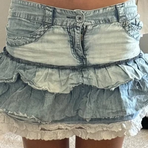 Volangig jeanskjol med spetskant - Supersöt kort jeanskjol med flera volanger och ljus spetskant längst ner. Kjolen har klassiska jeansdetaljer som fickor och dragkedja framtill. Perfekt för dig som gillar en lekfull och trendig stil. Volang handsydd och därav pris, SÄNKER INTE PRIS!