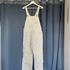 Randiga hängslebyxor från Carhartt WIP - Säljer en vit overall med svarta ränder från Carhartt WIP. Klassisk workwear-look med bröstficka, justerbara axelband och flera fickor. Materialet är en mix av bomull och polyester, vilket gör den både slitstark och bekväm. Perfekt för dig som gillar streetwear och utility vibes! ca 2000kr nypris
