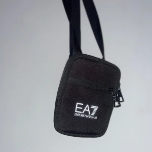 Svart axelväska EA7 Emporio Armani - Snygg svart axelväska från EA7 Emporio Armani med justerbar rem och dragkedja framtill. Väskan har stilren design med vit logotyp på framsidan och är tillverkad i slitstark polyester. Perfekt storlek för mobil, plånbok och andra småsaker.