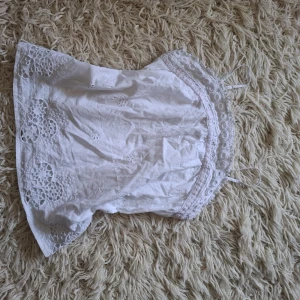 Vit broderad linnetopp med spets - Superfin vit linnetopp med tunna axelband och broderade blommor. Toppen har spetsdetaljer längs överkanten och små hålbroderier som ger en romantisk vibe. Perfekt för varma dagar och sommarhäng.