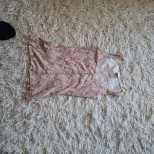 Blommig ribbad linnetopp i rosa och beige - Supersöt ärmlös linnetopp med blommigt mönster i rosa och beige. Toppen har ribbad struktur och knappar framtill som snygg detalj. Perfekt för varma dagar och enkel att matcha med jeans eller kjol.