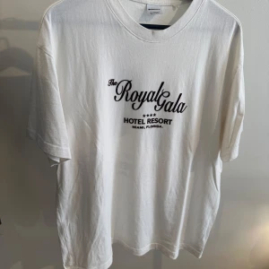 Vit t-shirt Royal Gala Hotel Resort - Vit t-shirt från Bershka med svart tryck fram och bak. På framsidan står det 'The Royal Gala Hotel Resort Miami, Florida' och på baksidan finns ett stort tryck med svarta loafers i sammet. Klassisk passform och rund hals. Perfekt för dig som gillar statement-plagg.