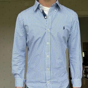 Blåvit randig skjorta från Polo Ralph Lauren - Klassisk skjorta från Polo Ralph Lauren i blåvitt randigt mönster och broderad logga på bröstet. Skjortan är köpt för 1800kr och näst intill aldrig använd. Skjortan är i storlek S och är skräddarsydd, perfekt för dig som är runt 170cm. Hör av dig vid frågor eller funderingar!