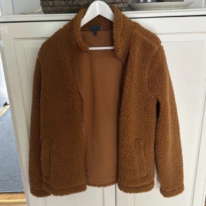 Brun teddyjacka från H&M - Mysig brun teddyjacka från H&M med regular fit. Jackan har dragkedja framtill, hög krage och två fickor. Perfekt för kyliga dagar när du vill ha en chill och trendig look. Fodrad insida och fluffig utsida som ger en skön vibe.