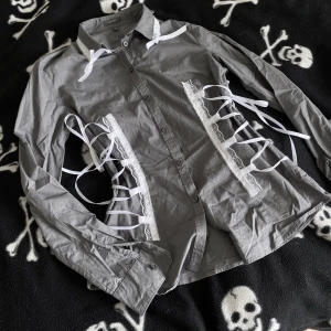 Grå handsydd skjorta med snörning och spets - Unik grå skjorta med vita snörningsdetaljer och spets längs sidorna och kragen. Långärmad modell i bomull med knappar framtill. Perfekt för dig som vill sticka ut med en edgy och kreativ look. Plagget är handsytt, vilket innebär att det kan förekomma mindre skavanker tex lösa trådar! 